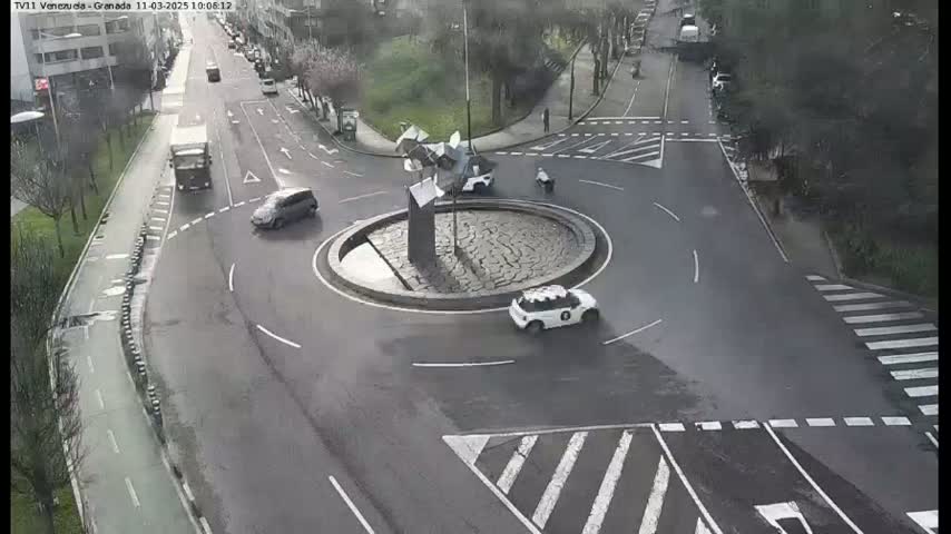 A wet roundabout with several vehicles and a sculpture in the center is surrounded by roads and pedestrian crossings.