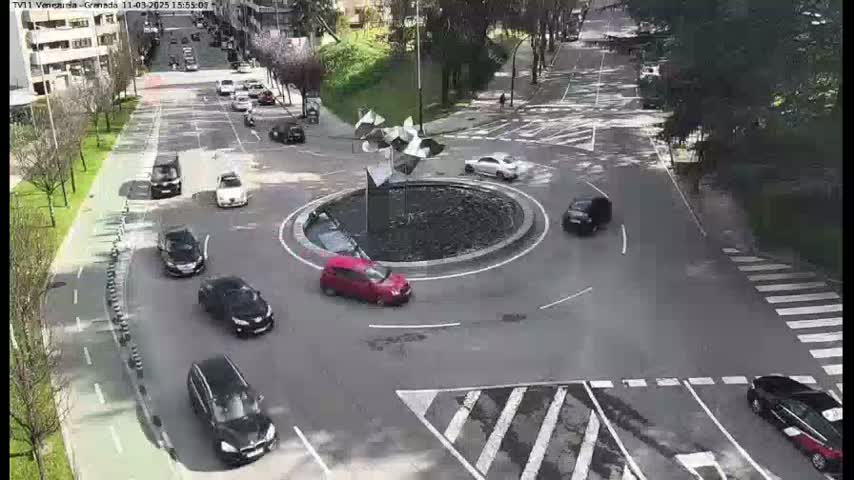 Several cars are driving around a traffic circle with a fountain in the center, on a mostly clear day.