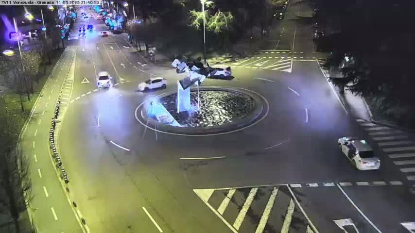 At night, several cars navigate a roundabout centered around a water feature, with streetlights illuminating the scene.