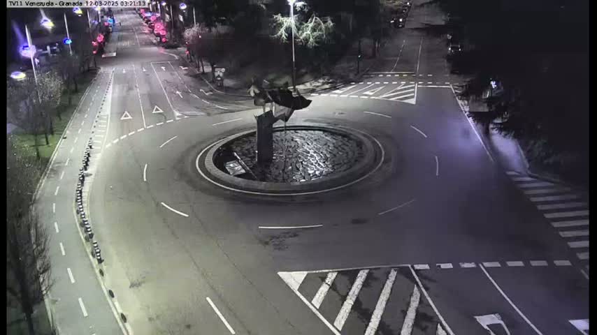 A nighttime aerial view of a roundabout with a dark sculpture in the center, surrounded by roadways with sparse traffic.