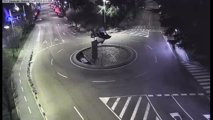 A nighttime aerial view of a roundabout with a sculpture in the center, surrounded by roads with crosswalks and parked cars along the sides.