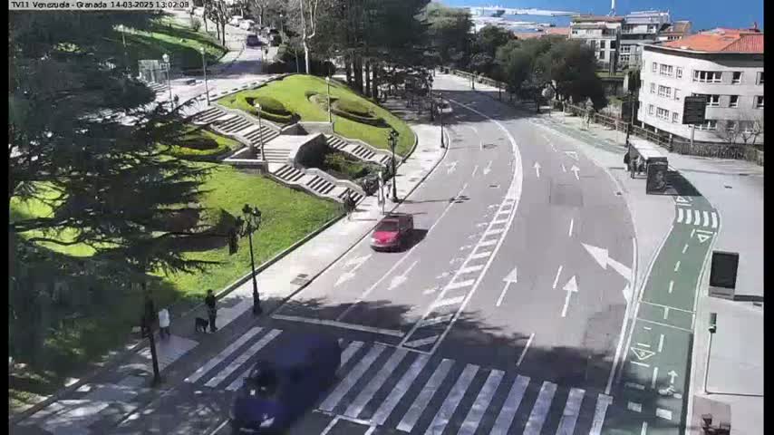 A sunny day overlooking a curved road with a pedestrian crossing, alongside a grassy park with stairs and a few people walking.