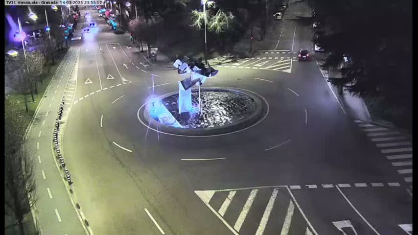 A nighttime aerial view shows a roundabout with a modern sculpture in the center, surrounded by roadways with minimal traffic.