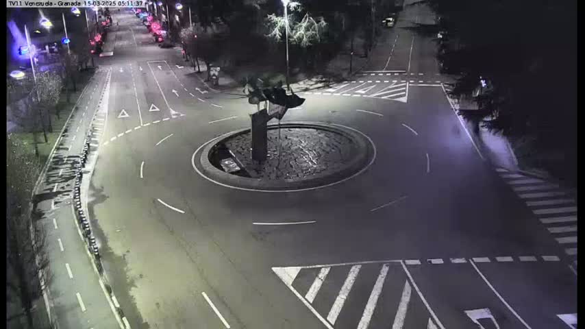 A nighttime aerial view of a roundabout with a dark, abstract sculpture in the center, surrounded by roadways with minimal traffic.