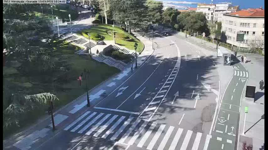 A sunny day reveals a paved roadway with crosswalks and bike lanes curving through a park-like setting with stairs, greenery, and a few buildings in the background.