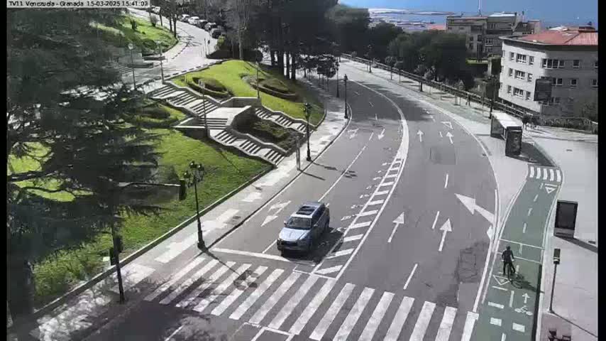 A sunny day shows a curving road with a car and cyclist, passing by landscaped areas with stairs, trees, and buildings in the background.