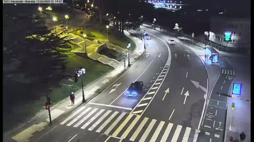 A nighttime street scene shows a car driving on a curved road that passes a pedestrian crossing and stairs leading up to a building, with several streetlights illuminating the area.