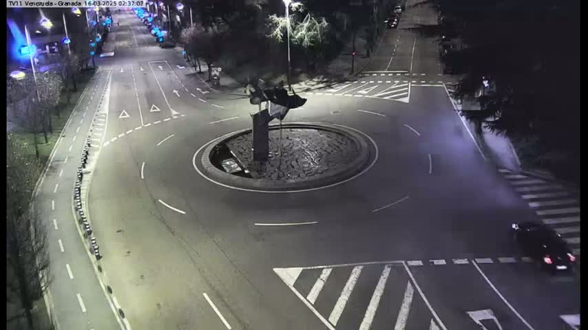 A nighttime aerial view shows a roundabout with a dark sculpture in the center, surrounded by roads with minimal traffic.