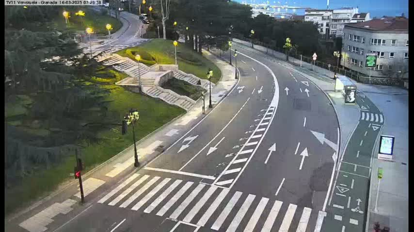 A curving roadway with pedestrian crosswalks and a bike lane is seen at dusk, next to a grassy area with stairs and some trees.