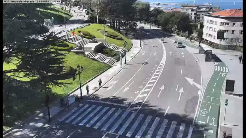 A sunny day reveals a curving road with a pedestrian crossing, flanked by a grassy park area with stairs and a few people walking.