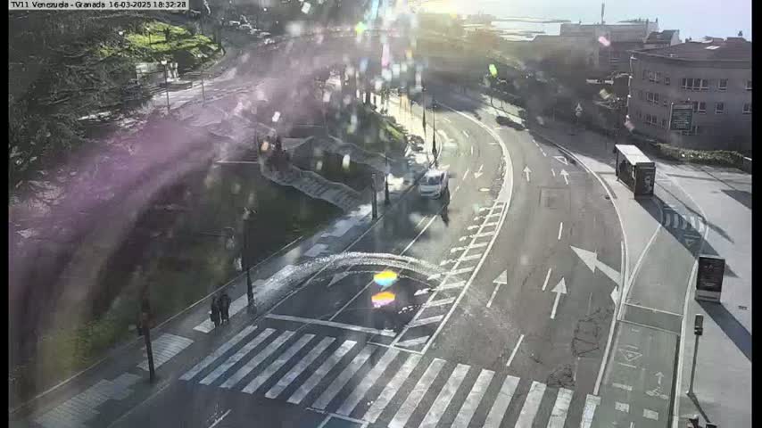 A sunny overhead view shows a road curving around a pedestrian crossing with a few people walking and a vehicle driving, near a bus stop and a building.