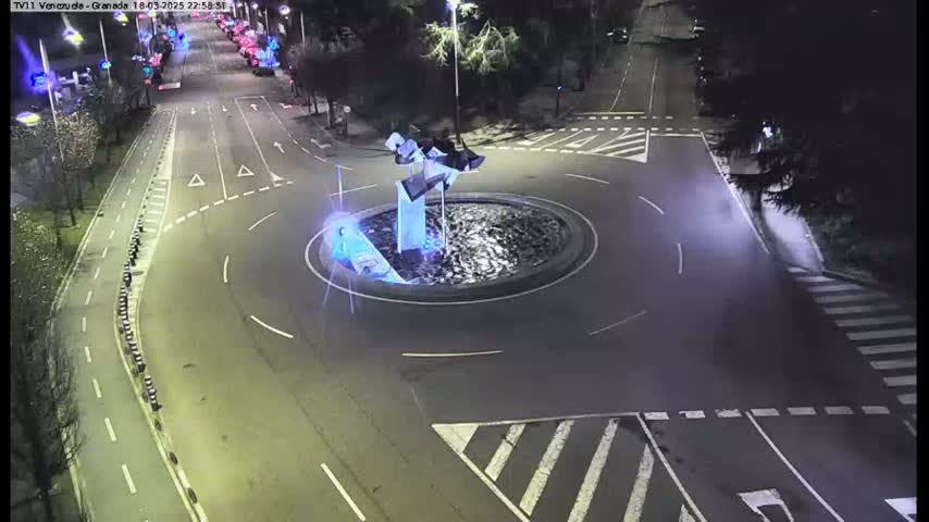 A nighttime aerial view shows a roundabout with a modern sculpture in the center, surrounded by roads and pedestrian crossings.