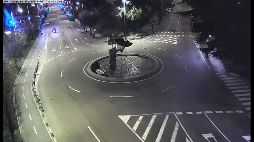A nighttime overhead view shows a roundabout with a dark sculpture in the center, surrounded by roads and pedestrian crossings.