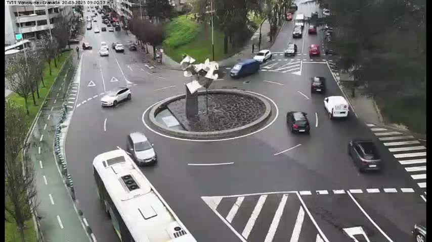 A wet roundabout with several cars and a bus, surrounded by streets with more vehicles.