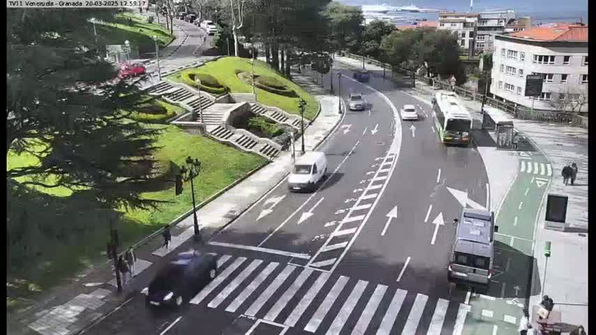 A sunny day shows a curved road with several cars and buses, a pedestrian crossing, and a grassy area with stairs.
