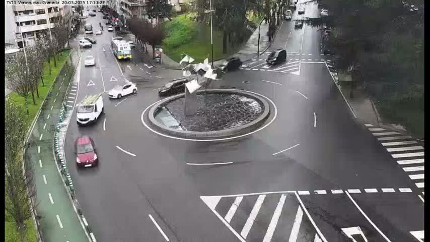 Several cars are navigating a wet roundabout with a modern sculpture in the center, surrounded by roads and pedestrian crosswalks.
