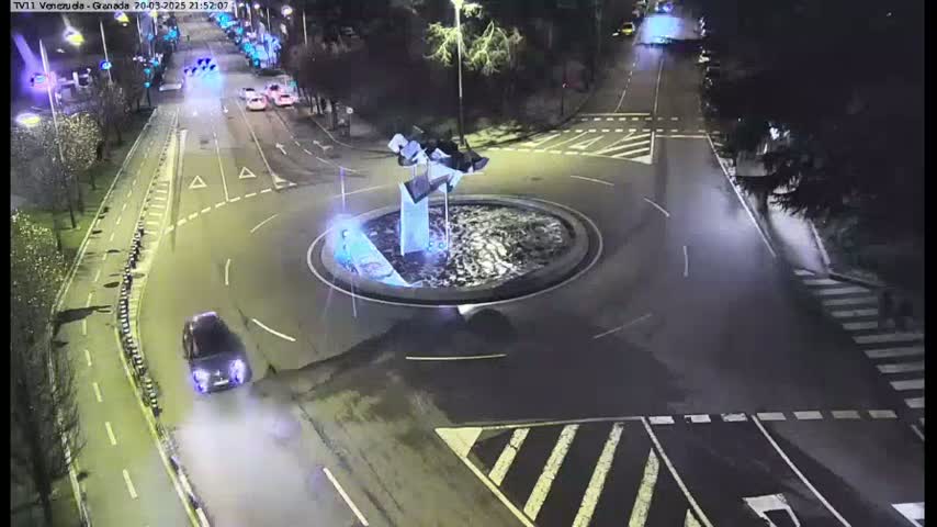 A nighttime aerial view shows a roundabout with a modern sculpture in the center, surrounded by roads with several vehicles, and a pedestrian crossing.