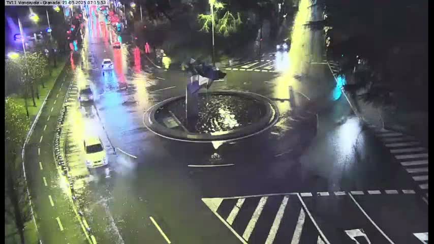 A wet, nighttime roundabout with a fountain in the center is surrounded by roadways with cars driving through it.