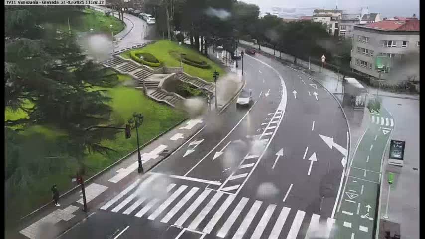 A wet road curves through a park-like setting, with a pedestrian crossing and a bike lane visible, under a rainy sky.