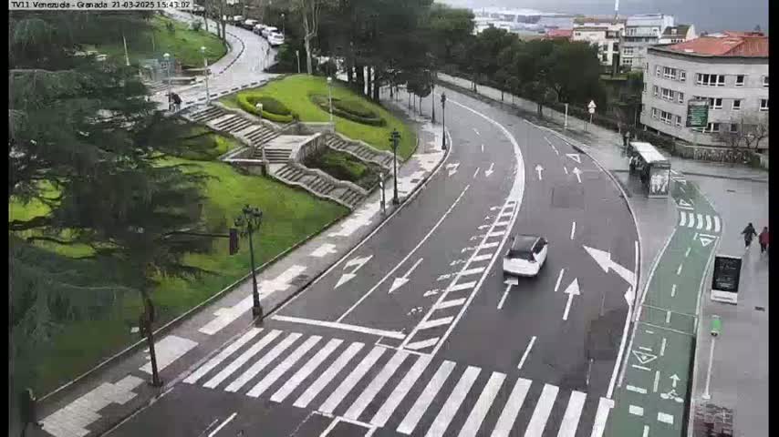 A wet, curving road with a crosswalk and bike lane winds through a grassy area with stairs and trees, under an overcast sky.