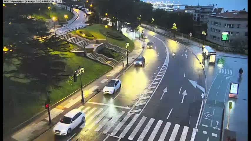 A wet, curving road with cars and pedestrians is flanked by stairs, green spaces, and buildings under dim, nighttime lighting.