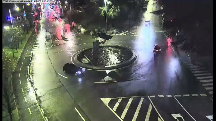 A wet, nighttime street scene shows several vehicles driving around a circular fountain.