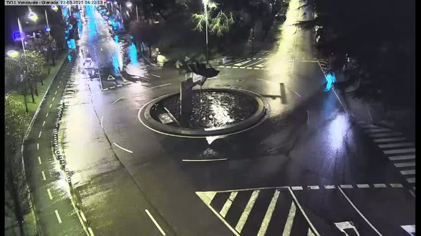 A wet, nighttime view of a roundabout with a sculpture in the center, surrounded by roadways and pedestrian crosswalks.