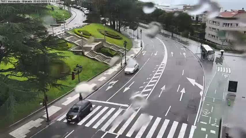 A rainy day scene shows a roadway with several cars and a bus,  a pedestrian crossing, and a staircase leading to a park-like area with green lawns.
