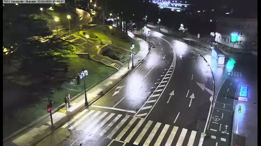 A wet, empty road curves through a nighttime city scene, passing a crosswalk where two people stand.