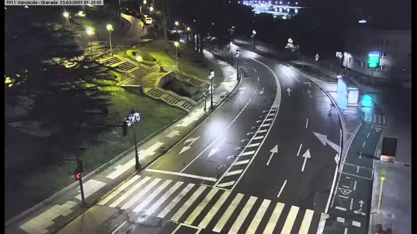 A wet, empty road curves through a nighttime scene, passing a pedestrian crossing near stairs leading up to a park-like area with trees and lights.