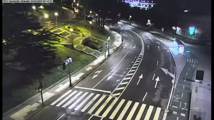 A mostly empty, well-lit roadway curves through a nighttime scene with pedestrian crosswalks, stairs leading up a grassy hill, and some buildings in the distance.