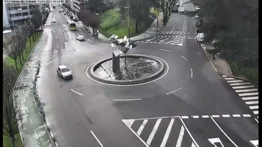 A wet roundabout with a modern sculpture in the center is surrounded by roads and crosswalks.