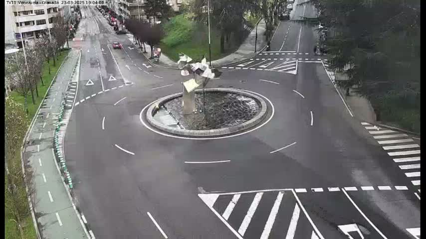 A wet roundabout with a modern sculpture in the center is surrounded by roads with light traffic.