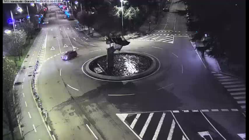 A dark, nighttime scene shows a roundabout with a single car in it and surrounding roads, with streetlights illuminating the area.