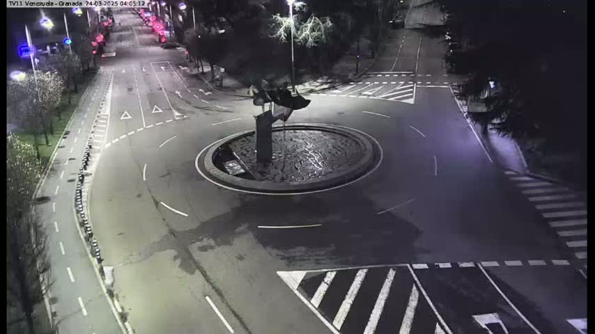 An overhead nighttime view of a traffic circle with a dark, abstract sculpture in the center, surrounded by wet asphalt roads and pedestrian crossings.
