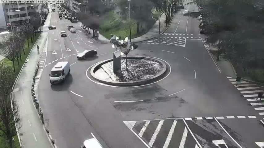 A wet roundabout with a modern metal sculpture in the center, surrounded by city streets with sparse traffic.