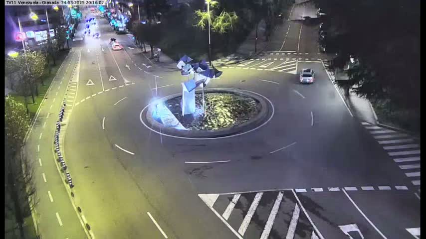 A nighttime overhead view shows a roundabout with a modern sculpture in the center, surrounded by roadways with cars and pedestrian crossings.