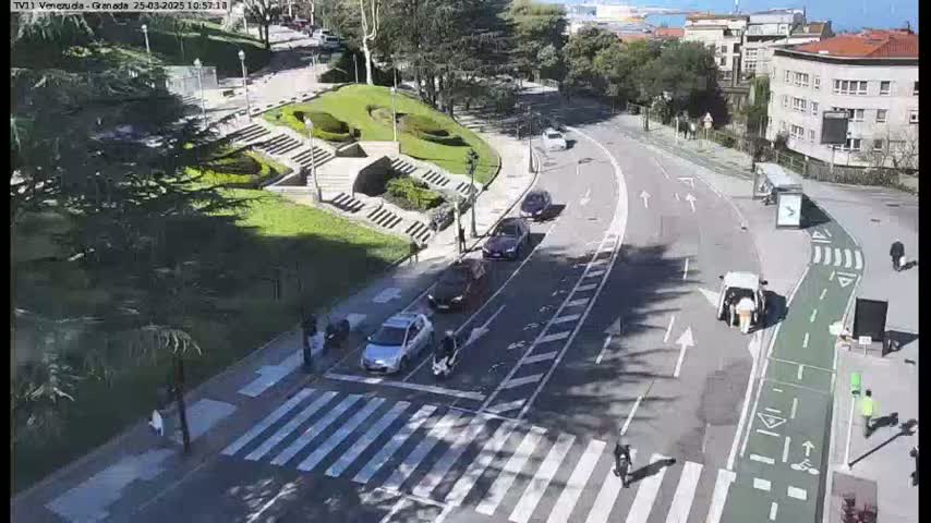 A sunny day shows a street scene with cars and pedestrians near a park with stairs and landscaping.