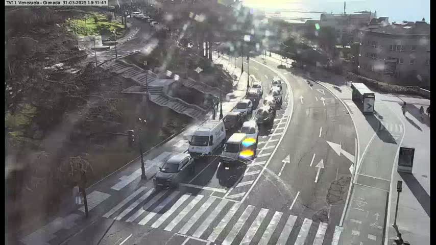A sunny day reveals a curving road with a line of cars, a van, and a bus, all near a pedestrian crossing and stairway leading down to a waterfront.