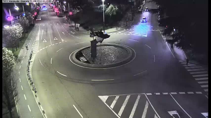 A nighttime aerial view of a roundabout with a sculpture in the center, surrounded by roads and pedestrian crossings.