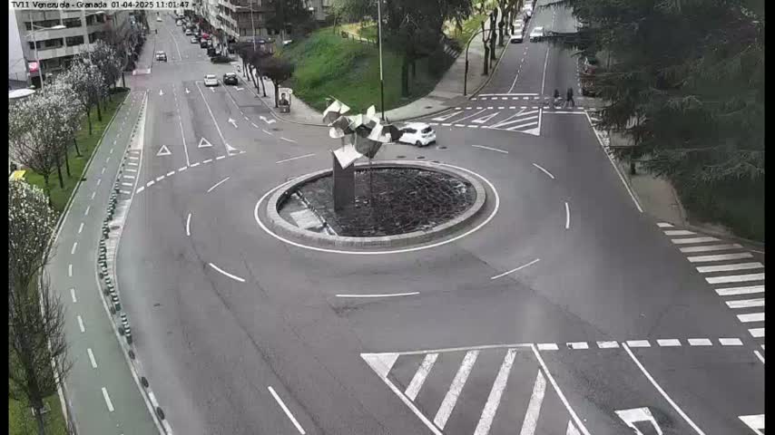 A mostly empty roundabout with a modern sculpture in the center is surrounded by roads with crosswalks and some cars and pedestrians.