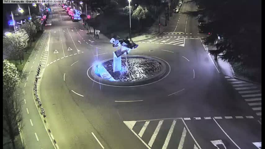 A nighttime aerial view of a roundabout with a modern sculpture in the center, surrounded by roads and pedestrian crossings.