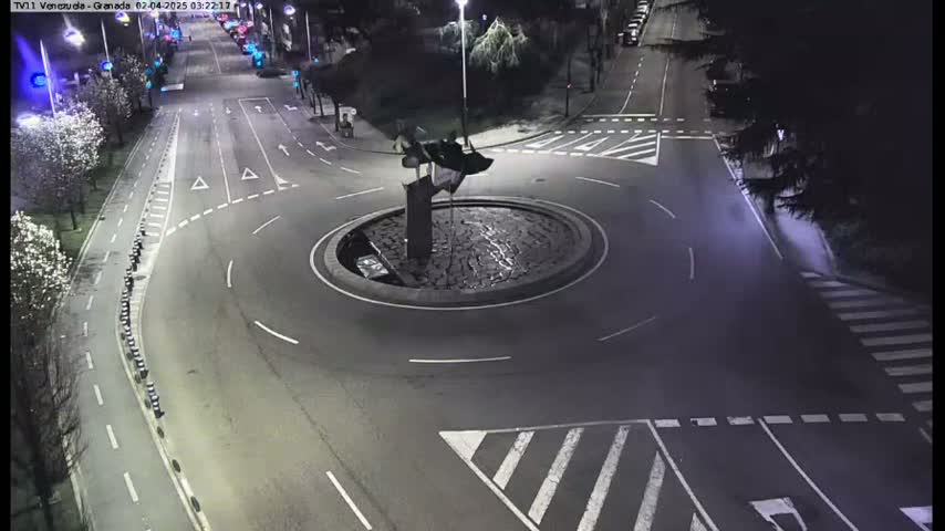 A nighttime aerial view shows a roundabout with a dark sculpture in the center, surrounded by roads with white markings and sparse traffic.