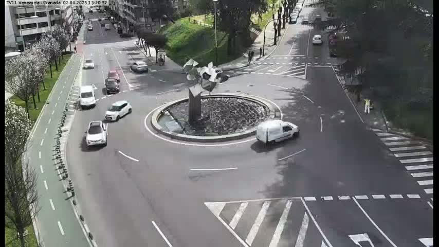 A roundabout with a fountain in the center is surrounded by cars and a pedestrian, under a mostly sunny sky.