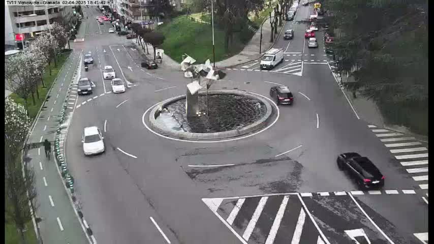 An aerial view shows a traffic roundabout with several cars driving around it and a modern sculpture in the center, on a partly cloudy day.