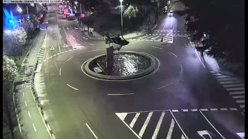 A nighttime aerial view of a roundabout with a dark statue in the center, surrounded by roads and crosswalks.