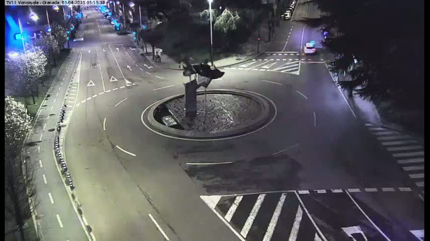 A nighttime aerial view shows a roundabout with a dark sculpture in the center, surrounded by roads and pedestrian crossings.