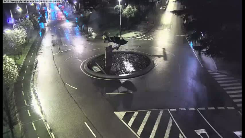 A wet roundabout at night with a sculpture in the center, surrounded by roadways and pedestrian crossings.