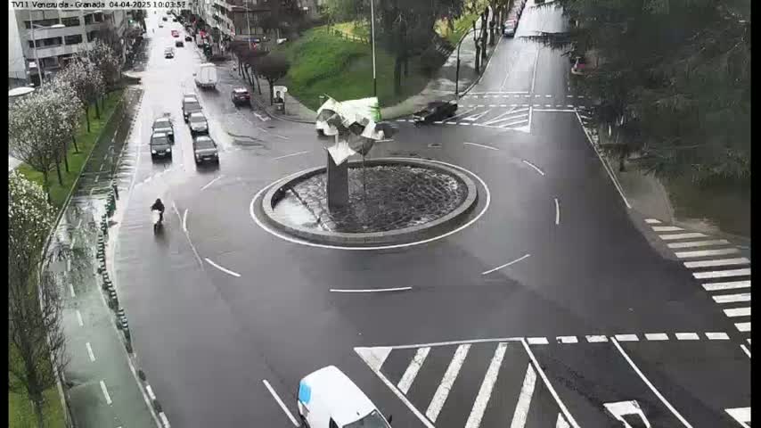 A wet roundabout with several cars and a person on a scooter is surrounded by roads and trees on a rainy day.