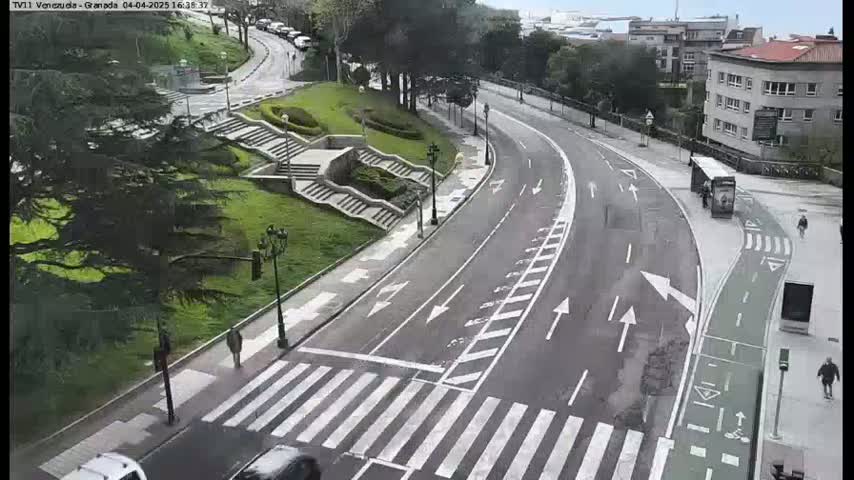 A paved road curves through a park-like setting with pedestrian crosswalks, a bike lane, and stairs leading up a grassy hill, under an overcast sky.